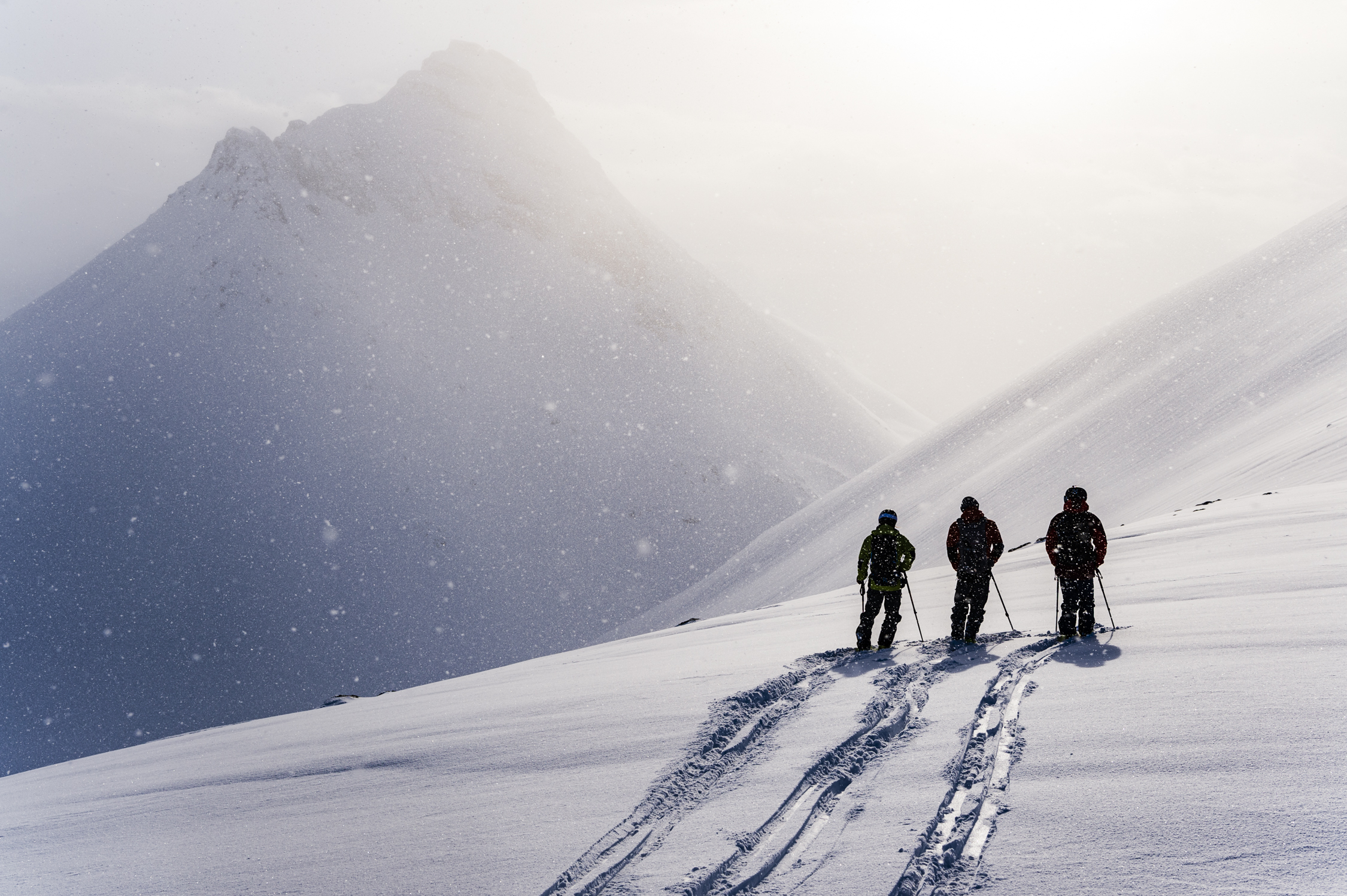 Siluetter av tre stillastående skidåkare mot en disig solnedgång mellan två fjäll.